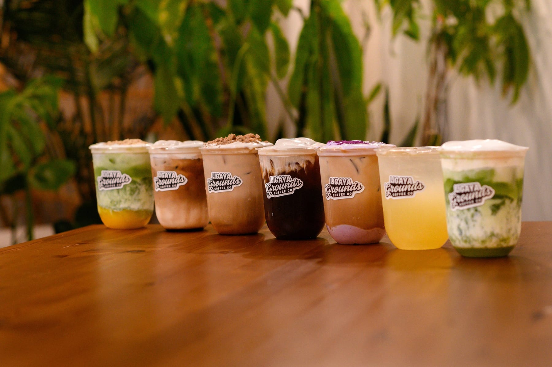 Seven Ligaya Grounds drinks lined up on a wooden table — matcha, iced lattes, cold brew, ube and calamansi — each in a branded cup, with palms behind.